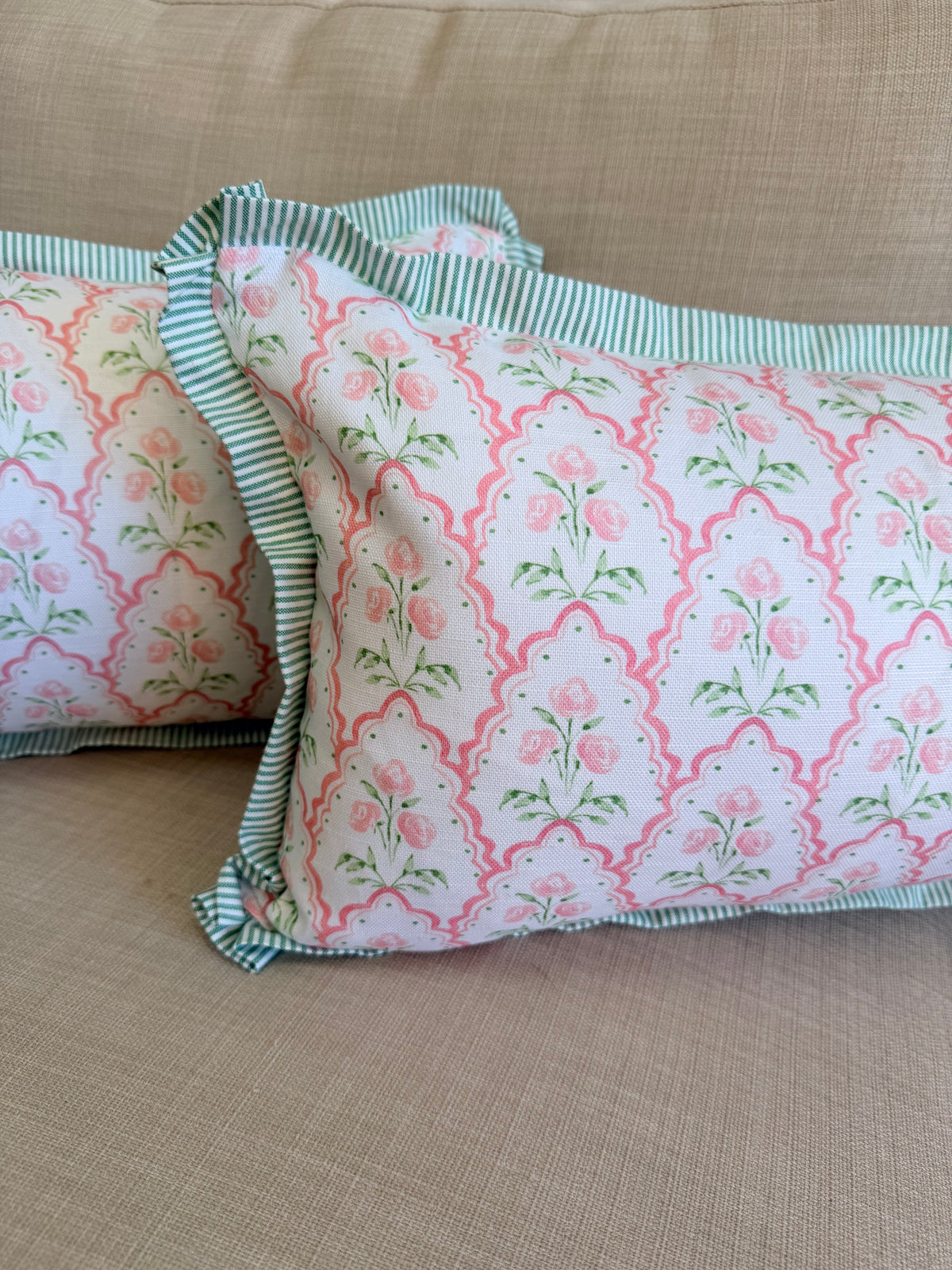 Two decorative throw pillows with a pink and green floral trellis pattern and striped green ruffled trim, resting on a beige textured fabric sofa.