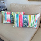 Two decorative throw pillows with colorful vertical stripes and blue geometric border panels, resting on a beige fabric sofa in a living room setting.