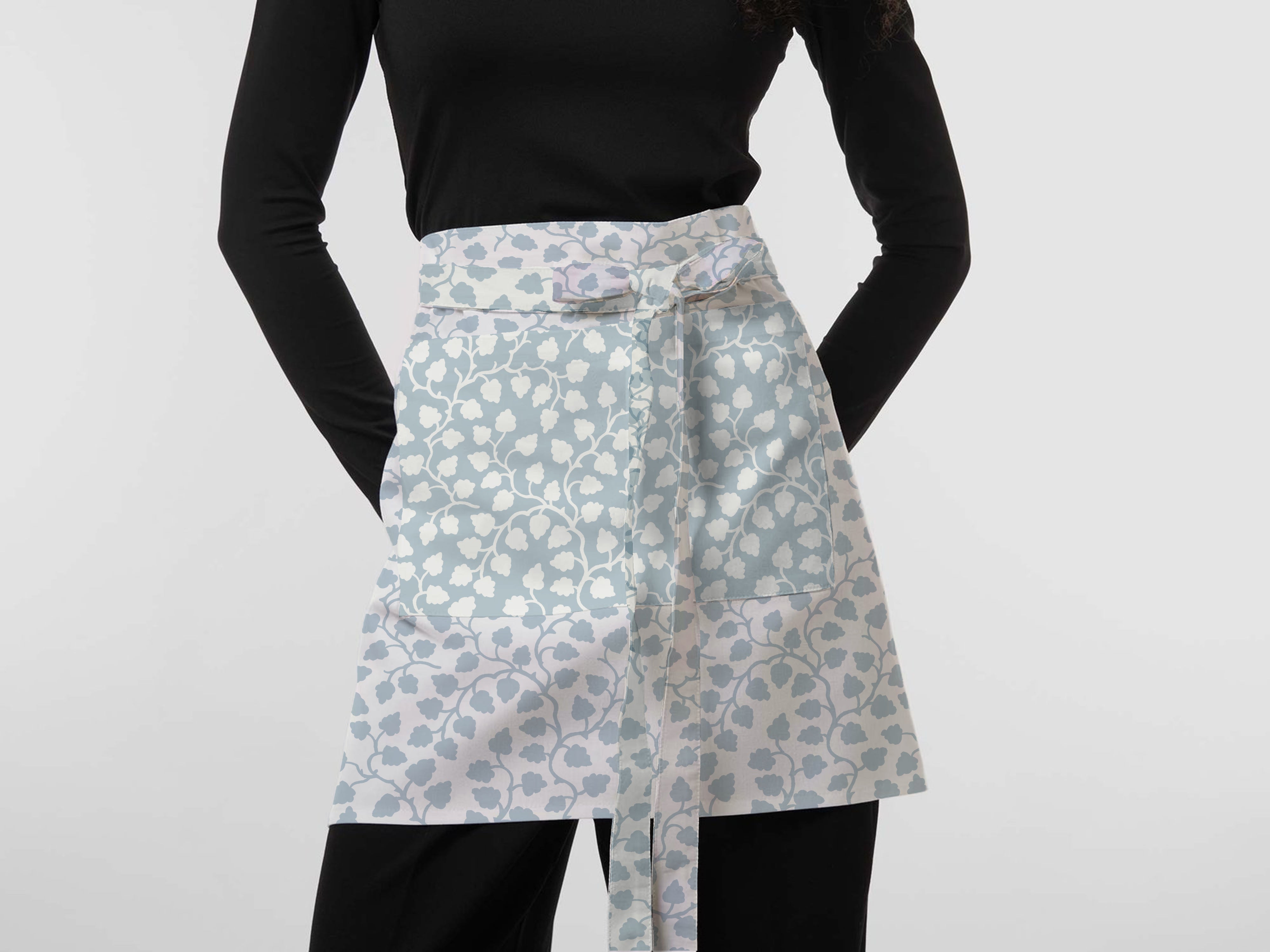 A model wearing a light blue and white floral patterned apron tied at the waist, paired with a black long-sleeve top and black pants, against a plain light gray background.