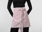 A model wearing a pink and white floral patterned apron tied at the waist over a black long-sleeve top and black pants, against a plain light gray background.