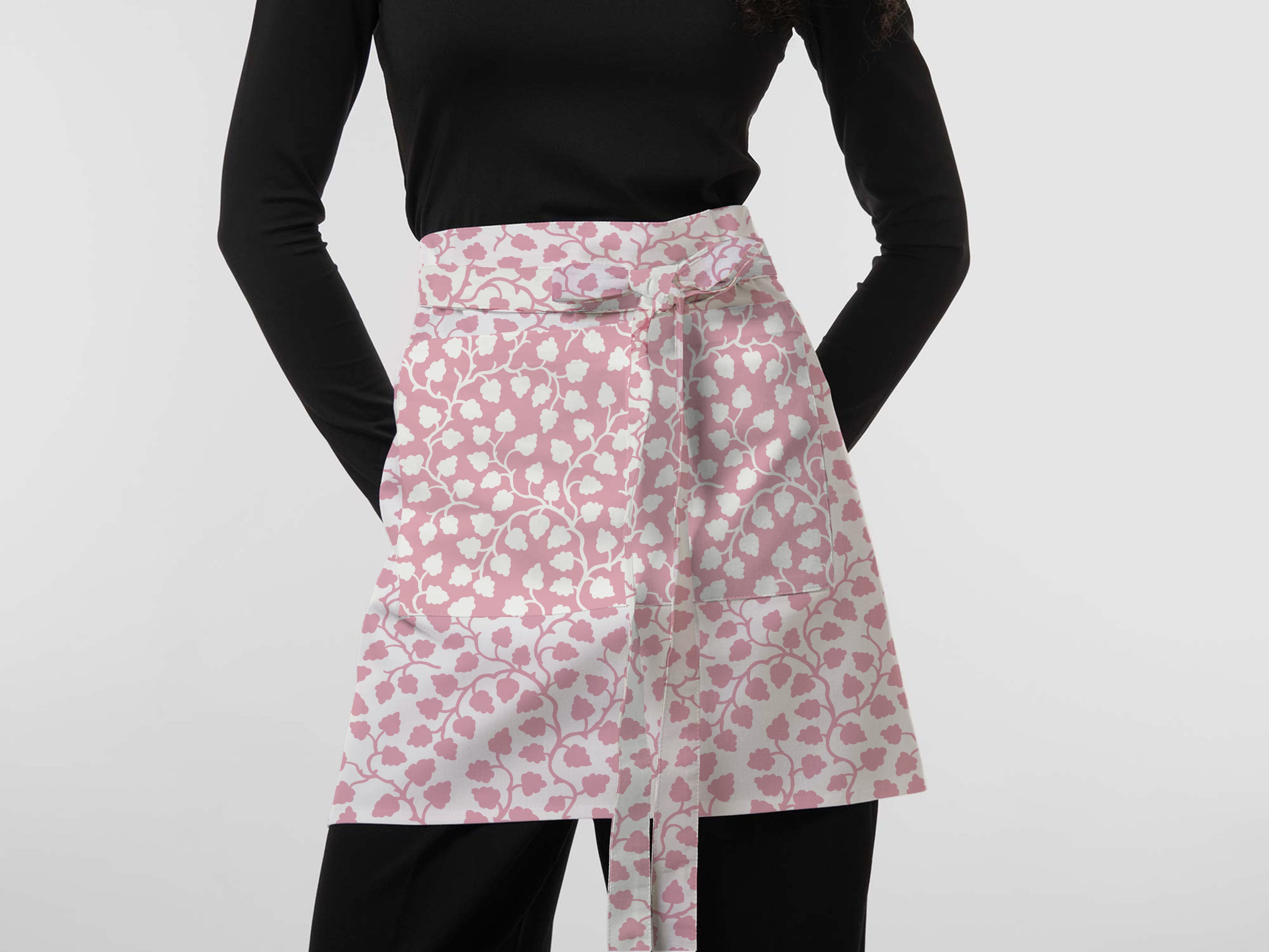 A model wearing a pink and white floral patterned apron tied at the waist over a black long-sleeve top and black pants, against a plain light gray background.